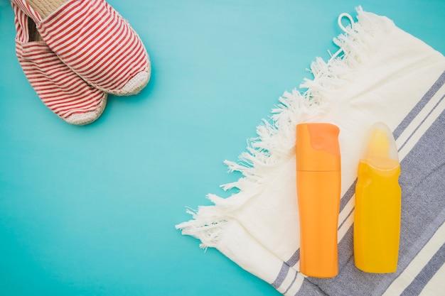 Summer beach essentials including a pair of striped red and white sandals, a white towel with fringe, and two bottles of sunscreen on a bright turquoise background.