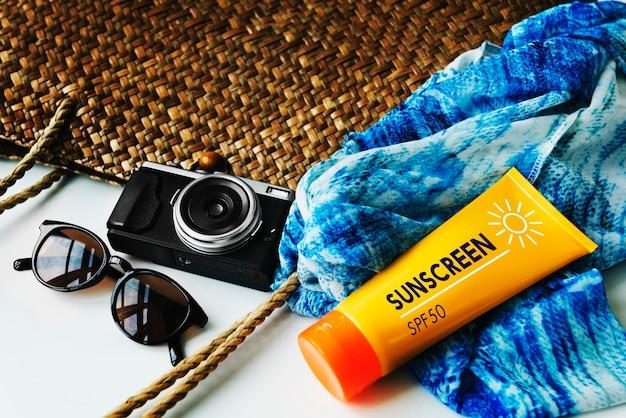 Summer travel essentials on a woven background, including sunglasses, an orange bottle of sunscreen SPF 50, a blue scarf, and a vintage camera.