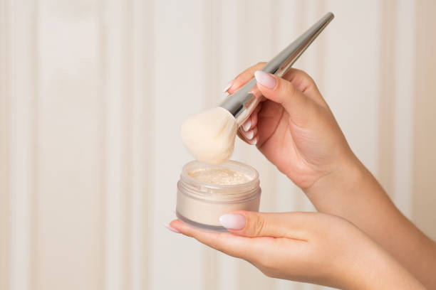 Close-up of a woman's hand applying loose powder with a brush from a small container.